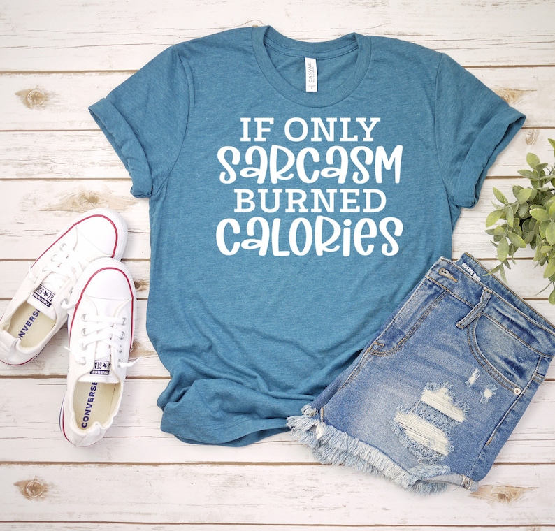 May include: A teal blue t-shirt with the text "If only sarcasm burned calories" in white lettering. The shirt is laid out on a wooden surface with a pair of white trainers and denim shorts.