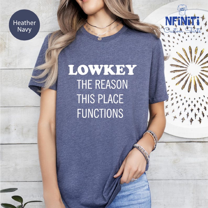 A woman wearing a blue t-shirt with the text "LOWKEY THE REASON THIS PLACE FUNCTIONS" printed on it.