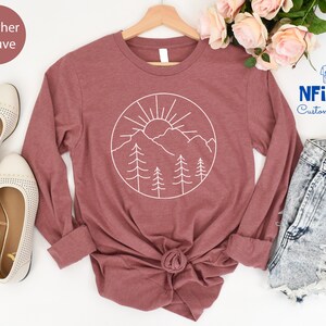 K&ouml;nnte beinhalten: Langarm-T-Shirt in Heather Mauve mit einem wei&szlig;en Umriss einer Sonne, Bergen und B&auml;umen in einem Kreis. Das Shirt liegt flach auf einer wei&szlig;en Oberfl&auml;che mit einem Paar wei&szlig;en Schuhen und einer Jeans-Shorts.