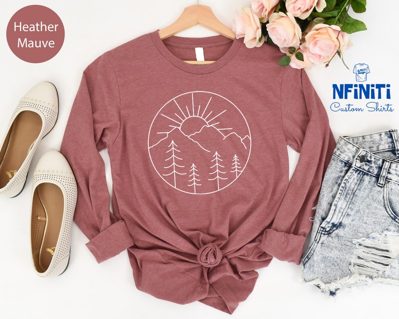K&ouml;nnte beinhalten: Langarm-T-Shirt in Heather Mauve mit einem wei&szlig;en Umriss einer Sonne, Bergen und B&auml;umen in einem Kreis. Das Shirt liegt flach auf einer wei&szlig;en Oberfl&auml;che mit einem Paar wei&szlig;en Schuhen und einer Jeans-Shorts.