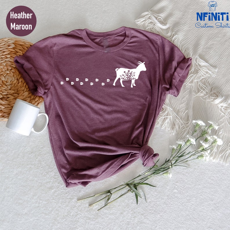 Pu&ograve; includere: Una t-shirt color marrone rossiccio con una silhouette bianca di una capra con dettagli floreali e una scia di impronte di zoccoli dietro.