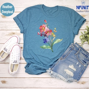 May include: A teal blue t-shirt featuring a watercolour floral design with pink, red, blue and yellow flowers. The shirt is lying on a white wooden surface with a pair of white trainers and denim shorts.