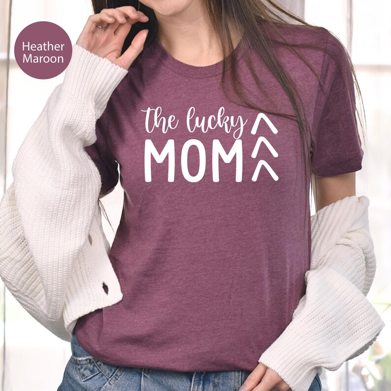 May include: Heather maroon t-shirt with the text "the lucky MOM" in white. The shirt has short sleeves and a crew neck. The text is in a handwritten style font. The shirt is made of a soft material.