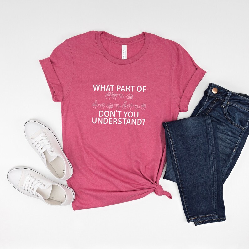 May include: A pink t-shirt with the text "WHAT PART OF DON'T YOU UNDERSTAND?" in white. The text is accompanied by a graphic of the American Sign Language alphabet for the word "deaf".