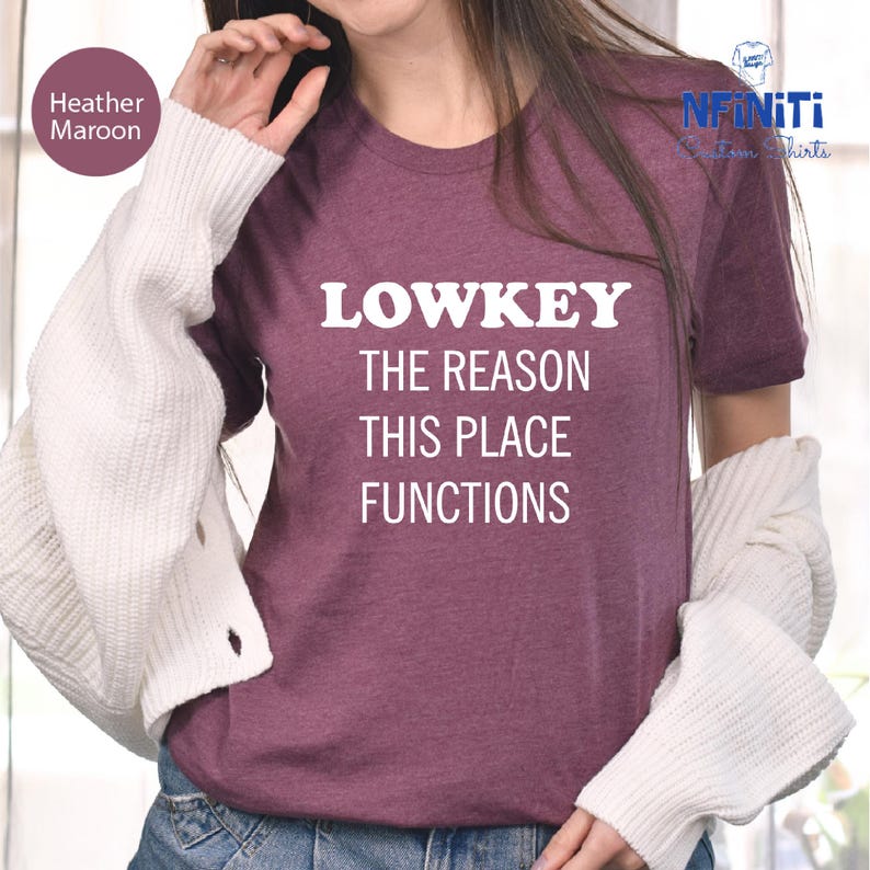 A person wearing a purple t-shirt with the text "LOWKEY THE REASON THIS PLACE FUNCTIONS" printed on it.
