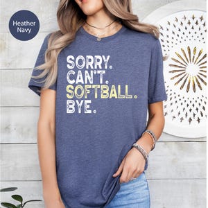 May include: A heather navy short-sleeved t-shirt featuring the white and yellow distressed text SORRY. CAN'T. SOFTBALL. BYE. The shirt has a crew neckline and a standard fit, showcasing a casual style with a clear graphic print on the front.