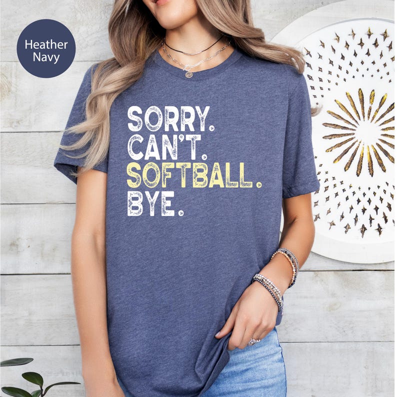 May include: A heather navy short-sleeved t-shirt featuring the white and yellow distressed text SORRY. CAN'T. SOFTBALL. BYE. The shirt has a crew neckline and a standard fit, showcasing a casual style with a clear graphic print on the front.