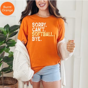 May include: A burnt orange short-sleeved t-shirt featuring white distressed text that reads SORRY. CAN'T. SOFTBALL. BYE. The shirt is worn with a white knit cardigan and light blue denim shorts, creating a casual look.