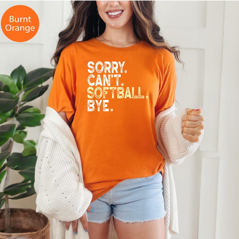 May include: A burnt orange short-sleeved t-shirt featuring white distressed text that reads SORRY. CAN'T. SOFTBALL. BYE. The shirt is worn with a white knit cardigan and light blue denim shorts, creating a casual look.