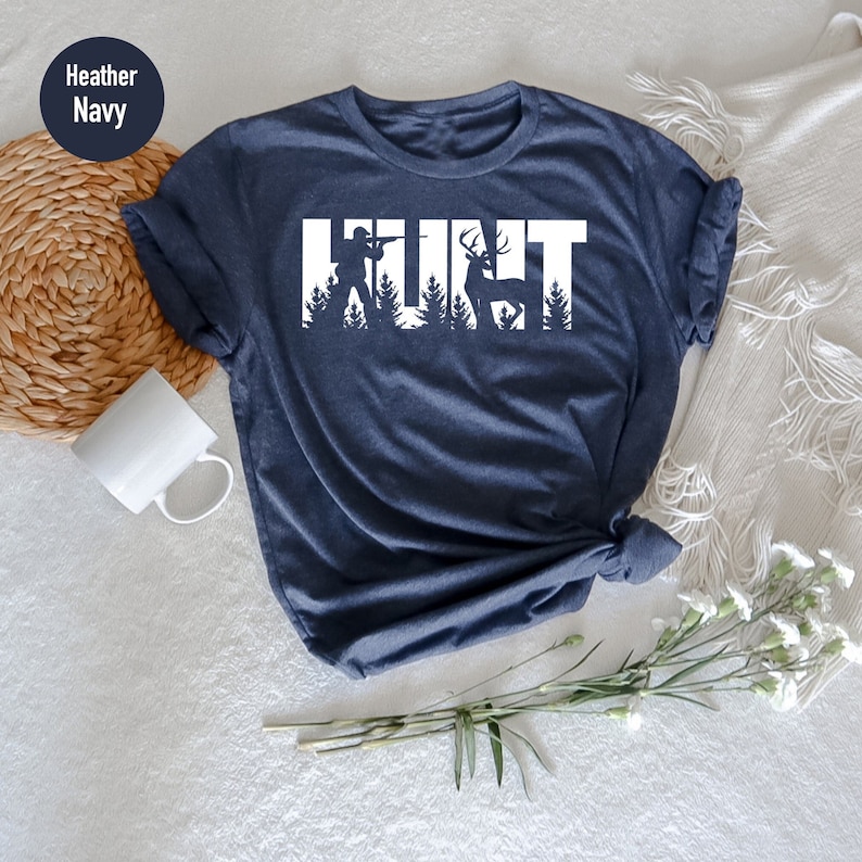 May include: A heather navy blue t-shirt with a white graphic of a hunter and deer with the word "HUNT" spelled out using the silhouettes of the hunter, deer, and trees.