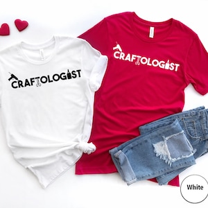 May include: Two t-shirts with the text "CRAFTOLOGIST" printed on them. One shirt is white with black text and the other is red with white text. Both shirts have a graphic of a glue gun, scissors, and a paint bottle.