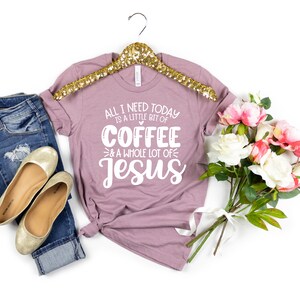 May include: Mauve t-shirt with the text "ALL I NEED TODAY IS A LITTLE BIT OF COFFEE & A WHOLE LOT OF Jesus". The shirt is paired with blue jeans, gold flats, and a bouquet of pink and white flowers. The shirt is hanging on a gold hanger.