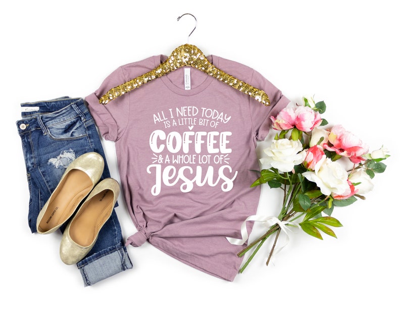 May include: Mauve t-shirt with the text "ALL I NEED TODAY IS A LITTLE BIT OF COFFEE & A WHOLE LOT OF Jesus". The shirt is paired with blue jeans, gold flats, and a bouquet of pink and white flowers. The shirt is hanging on a gold hanger.