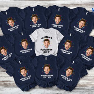 May include: A collection of navy blue and white t-shirts. Most shirts feature a smiling face graphic with the text "STRONGER WITH EVERY STEP." One white shirt reads "ALLISON'S MARATHON CREW."