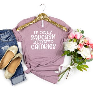 May include: A mauve t-shirt with the text "If only sarcasm burned calories" in white lettering. The t-shirt is on a gold hanger and is surrounded by a pair of jeans, gold flats, and a bouquet of white and pink flowers.
