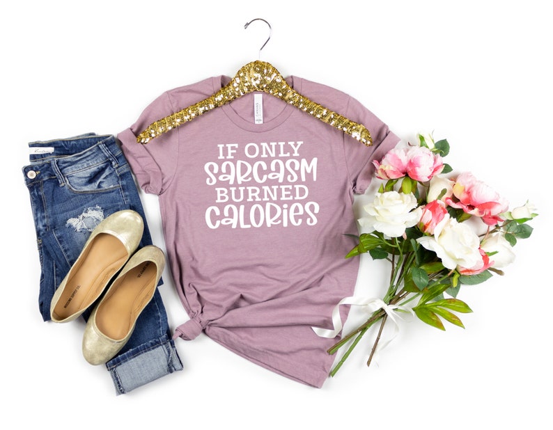 May include: A mauve t-shirt with the text "If only sarcasm burned calories" in white lettering. The t-shirt is on a gold hanger and is surrounded by a pair of jeans, gold flats, and a bouquet of white and pink flowers.