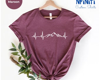 Könnte beinhalten: Heather-marronfarbenes T-Shirt mit einem weißen Bergpanorama und einem Herzschlag-Motiv. Das Shirt hängt an einem Kleiderbügel und hat einen weißen Hintergrund.
