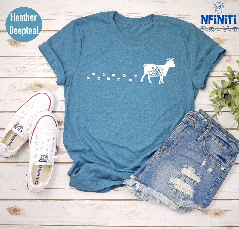 Pu&ograve; includere: Una t-shirt color blu profondo melange con un design di capra floreale bianca e il testo "NFINITI Custom Shirts". La t-shirt &egrave; appoggiata su una superficie di legno con un paio di scarpe da ginnastica bianche e un paio di pantaloncini di jeans.