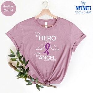 May include: Heather orchid-colored t-shirt with the text "my HERO is now my ANGEL" and a purple ribbon with wings, promoting Alzheimer's awareness. The shirt is displayed on a wooden hanger.