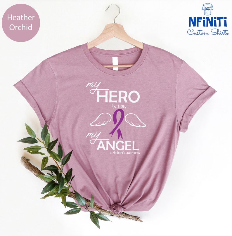 May include: Heather orchid-colored t-shirt with the text "my HERO is now my ANGEL" and a purple ribbon with wings, promoting Alzheimer's awareness. The shirt is displayed on a wooden hanger.