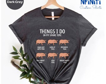 Könnte beinhalten: Dunkelgraues T-Shirt mit dem Text "Things I do in my spare time" und sechs Illustrationen von Schweinen mit verschiedenen Aktivitäten: mit Schweinen abhängen, Schweine betrachten, Schweine erforschen, über Schweine sprechen, Schweine füttern und von Schweinen träumen.