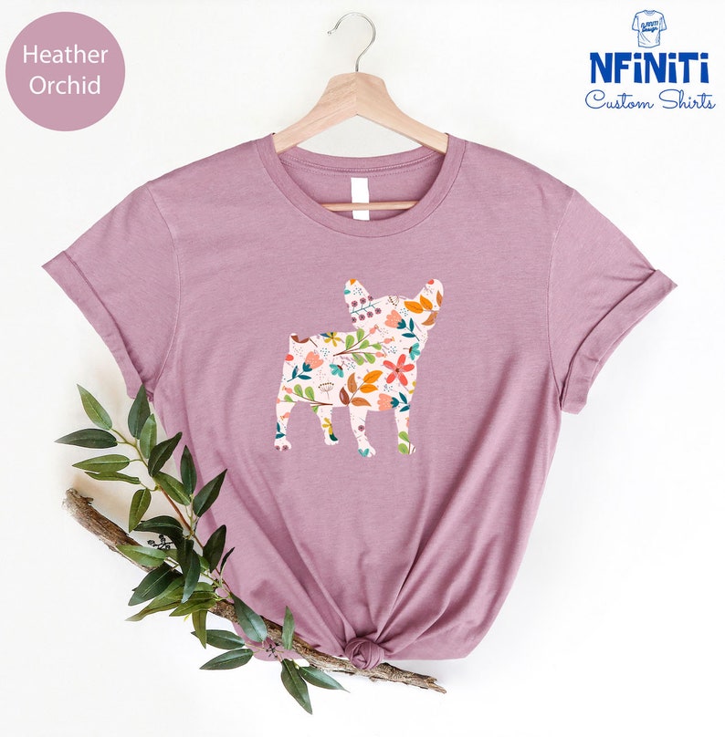 Peut inclure: Un t-shirt en heather orchid avec une silhouette de bouledogue fran&ccedil;ais &agrave; motif floral. Le t-shirt est sur un cintre et a un fond blanc.