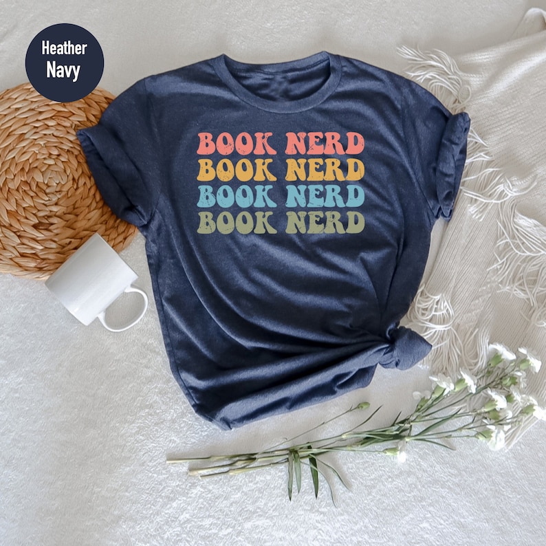 May include: A heather navy blue t-shirt with the text "BOOK NERD" repeated four times in a retro rainbow color scheme.