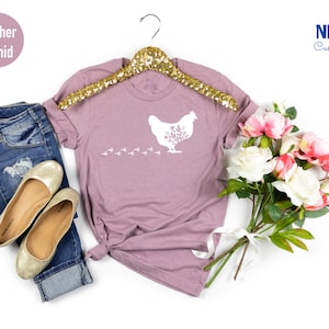 May include: A heather orchid t-shirt with a white silhouette of a chicken with flowers in its feathers and a trail of chicken footprints. The shirt is on a hanger and there is a bouquet of pink and white flowers in the background.