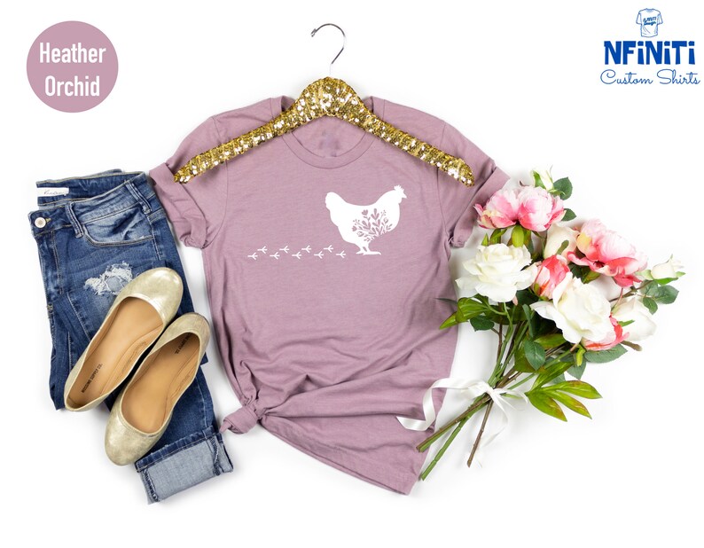 May include: A heather orchid t-shirt with a white silhouette of a chicken with flowers in its feathers and a trail of chicken footprints. The shirt is on a hanger and there is a bouquet of pink and white flowers in the background.