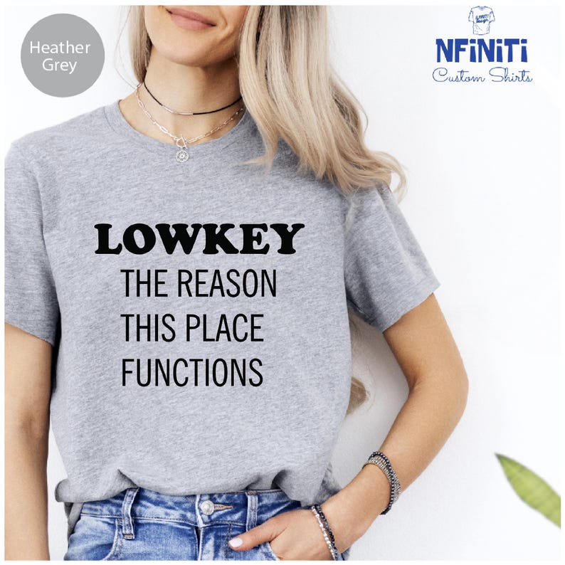 A woman wearing a grey t-shirt with the text "LOWKEY THE REASON THIS PLACE FUNCTIONS" printed on it.