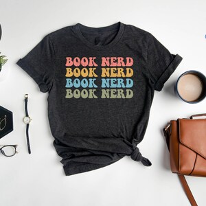 May include: A heather dark grey t-shirt with the text "BOOK NERD" repeated four times in rainbow colors. The text is in a retro font style.