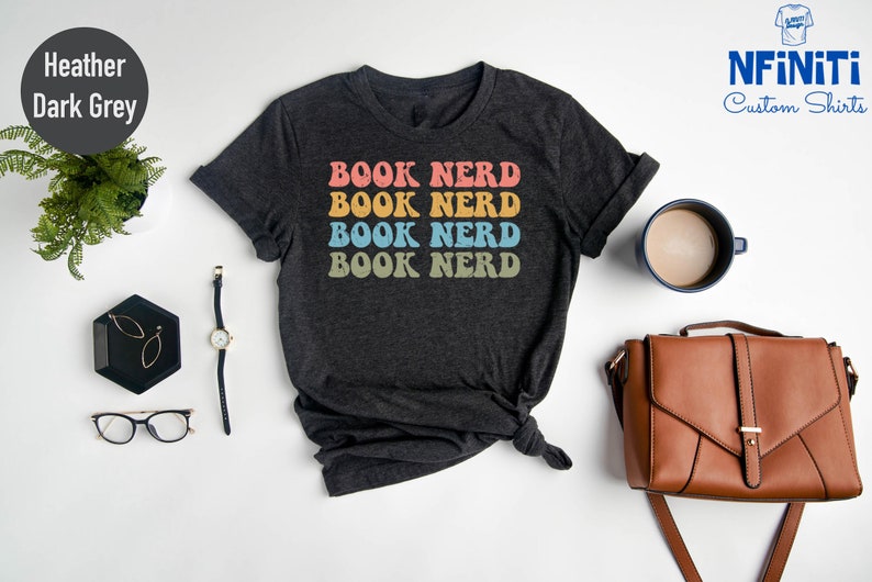 May include: A heather dark grey t-shirt with the text "BOOK NERD" repeated four times in rainbow colors. The text is in a retro font style.