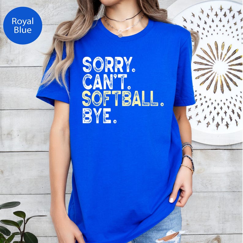May include: A royal blue short-sleeved t-shirt featuring white distressed text that reads SORRY. CAN'T. SOFTBALL. BYE. The shirt has a crew neckline and a straight hem, providing a standard fit for casual wear in a vibrant, solid colour.