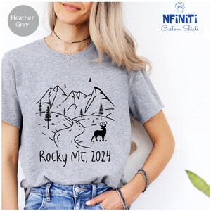 Peut inclure: T-shirt gris chin&eacute; avec un motif noir et blanc de montagnes, une route sinueuse, des arbres et un cerf. Le texte "Rocky Mt, 2024" est imprim&eacute; sous le motif.