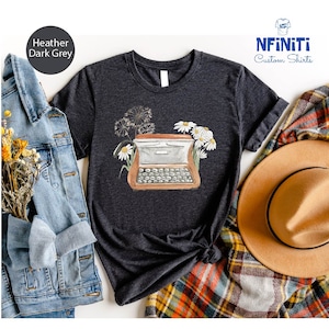 May include: A dark heather grey t-shirt with a vintage typewriter illustration and a floral design. The typewriter is brown with white keys and is surrounded by white daisies and a dandelion.