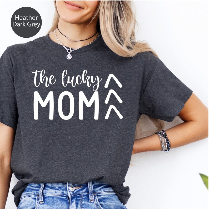 May include: Heather dark grey t-shirt with the text "the lucky MOM" in white script. The word "MOM" is below two upward-pointing arrows. The shirt is made of a soft material.