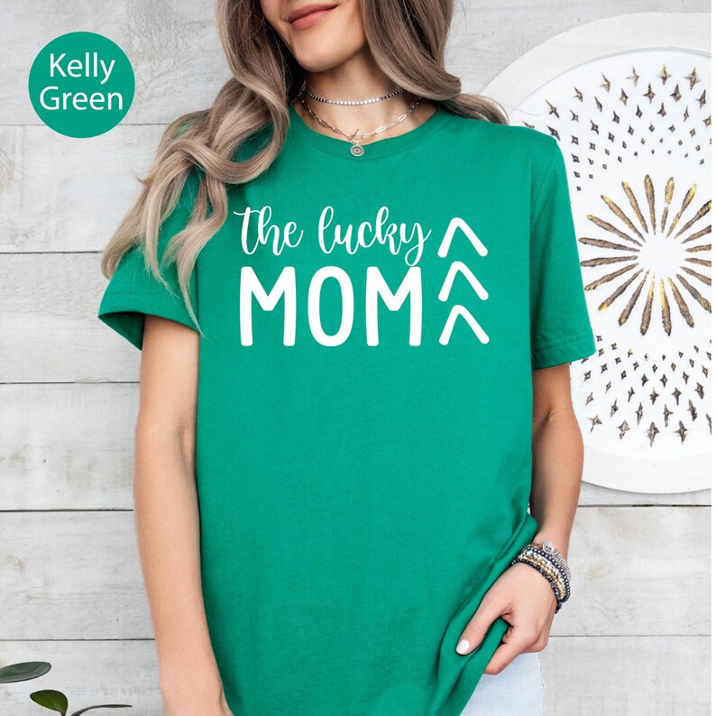 May include: A Kelly green t-shirt with the text "the lucky MOM" in white. The shirt has short sleeves and a crew neck. The words "Kelly Green" are in a green circle in the upper left corner.