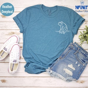 May include: A heather deepteal t-shirt with a white outline of a frog sitting on a branch. The shirt is laying on a white wooden surface with a pair of white converse shoes and a pair of denim shorts.