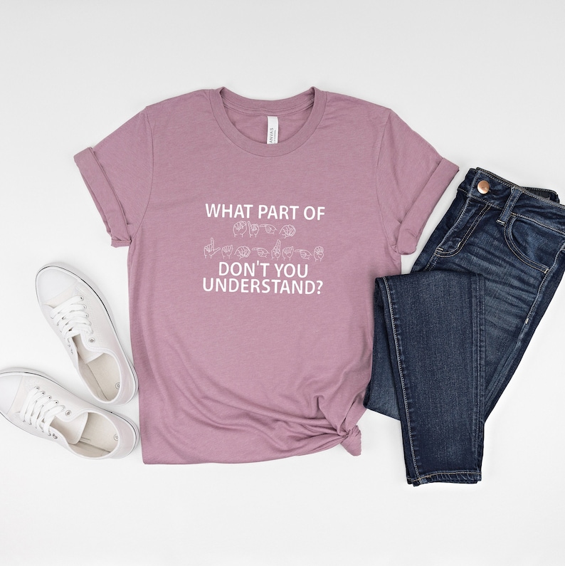 May include: A mauve t-shirt with the text "WHAT PART OF DON'T YOU UNDERSTAND?" in white lettering. The text is accompanied by a row of hand signs in American Sign Language.