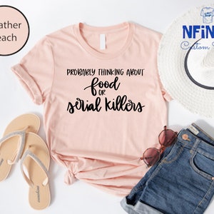 May include: A heather peach t-shirt with the text "Probably thinking about food or serial killers" in black.