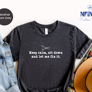 May include: A dark heather grey t-shirt with a white graphic of a pair of scissors and the text "Keep calm, sit down and let me fix it."