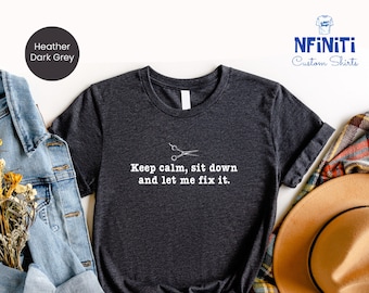 May include: A dark heather grey t-shirt with a white graphic of a pair of scissors and the text "Keep calm, sit down and let me fix it."
