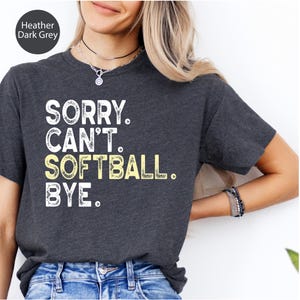 May include: Dark grey heather t-shirt featuring the text "SORRY. CAN'T. SOFTBALL. BYE." in white and yellow. The shirt has a crew neck.