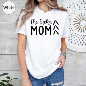 May include: White t-shirt with the text "the lucky MOM" in black. The shirt has a crew neck and short sleeves. The model is wearing blue jeans. The shirt is a casual style.