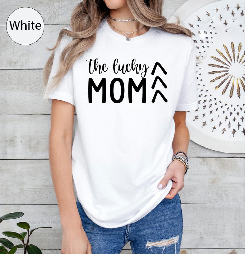 May include: White t-shirt with the text "the lucky MOM" in black. The shirt has a crew neck and short sleeves. The model is wearing blue jeans. The shirt is a casual style.