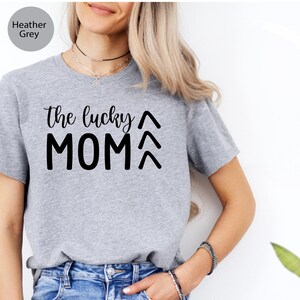 May include: Heather grey t-shirt with the text "the lucky MOM" in black script and block letters. Above and below the word "MOM" are two upward-pointing arrows. The shirt is made of a soft material and is suitable for casual wear.