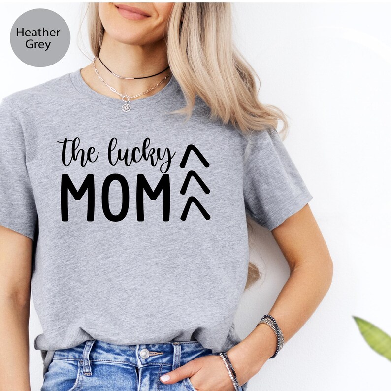 May include: Heather grey t-shirt with the text "the lucky MOM" in black script and block letters. Above and below the word "MOM" are two upward-pointing arrows. The shirt is made of a soft material and is suitable for casual wear.