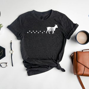 Pu&ograve; includere: Maglietta a collo a V color grigio scuro melange con una silhouette bianca di una capra che cammina con le impronte dietro di lei. La maglietta &egrave; stesa su una superficie bianca con una borsa di pelle marrone, una tazza di caff&egrave;, un orologio e un paio di occhiali.