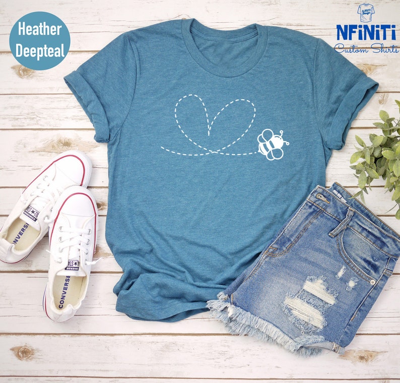 Peut inclure: Un t-shirt bleu sarcelle avec une abeille blanche volant en forme de c&oelig;ur compos&eacute; de lignes pointill&eacute;es. Le t-shirt est pos&eacute; sur une surface en bois blanc avec une paire de baskets blanches et un short en jean.