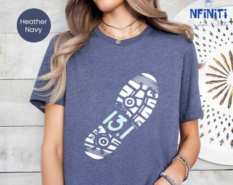 Könnte beinhalten: Ein heidefarbenes marineblaues T-Shirt mit einer weißen Grafik eines Fußabdrucks mit der Zahl "13" darin. Der Fußabdruck hat ein detailliertes Design eines Laufschuhprofils.